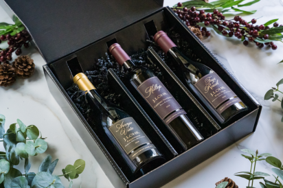 An open black box with three wine bottles places inside. The box is surrounded by decorative plants.