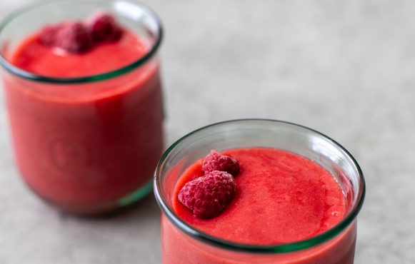 Image of two Raspberry Peach Smoothies. They are bright red, in glass containers, and topped with fresh raspberries.