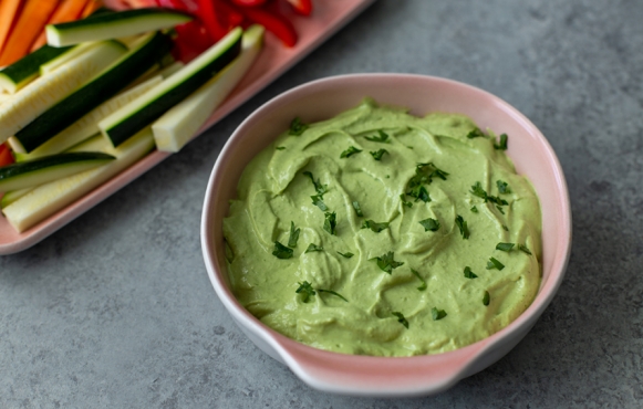 Avocado Cashew Dip next to some sliced vegetables. It is a green dip topped with fresh herbs.