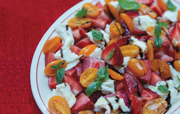 A caprese salad with mini colorful tomatoes, sliced strawberries, basil leaves and mozzarella cheese sit on a white plate on a red backdrop.